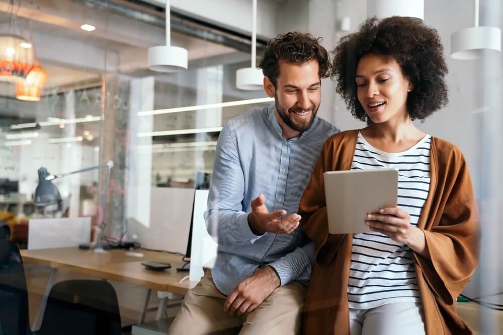 Two colleagues are in an office, with a man sitting on a desk and a woman holding a tablet. The man, with a beard and light blue shirt, listens intently as the woman, sporting curly hair, striped shirt, and brown cardigan, discusses DEI initiatives displayed on the tablet.